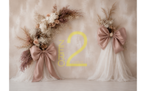 Baby girl photography backdrop with a textured beige wall, white draped fabric, botanical floral arrangements, and oversized dusty pink bows.