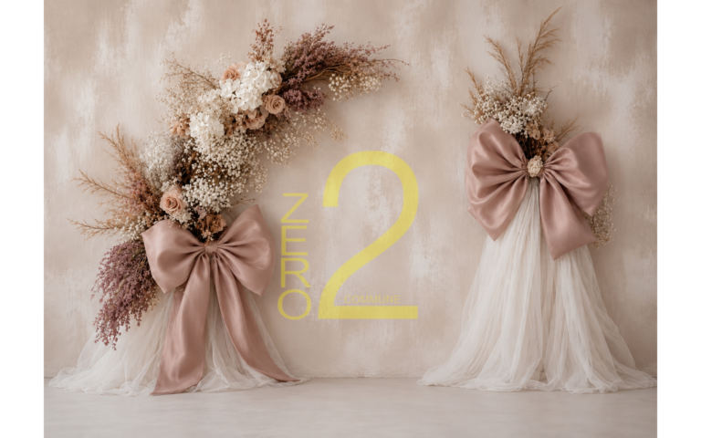 Baby girl photography backdrop with a textured beige wall, white draped fabric, botanical floral arrangements, and oversized dusty pink bows.
