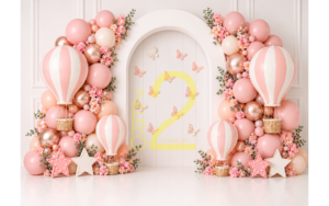 Pink hot air balloon baby girl birthday backdrop with pastel balloon clusters, butterflies, floral decorations, and a white arched panel.