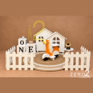 Outdoor theme baby photography setup with cream house props, orange scooter, white picket fence, ONE letter blocks, and woven round mat.