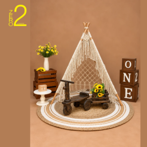 Outdoor-themed baby photography setup with macramé teepee tent, vintage wooden tricycle, wooden crates, flowers, and woven rug