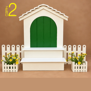 White cottage-style baby photography prop with green arched door, bench, white picket fence, and yellow flowers